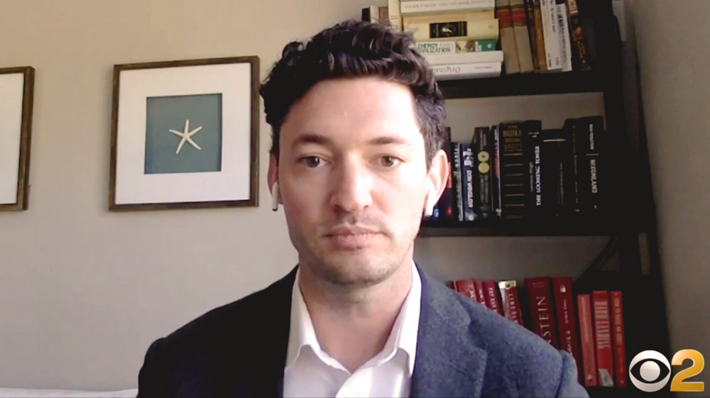 Andrew Buher in a suit facing a webcam with bookshelf behind him